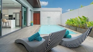 A modern outdoor pool area with lounge chairs and a kitchen visible through glass doors. - Nicky 3Br Private Pool Villa Bangtao (Choeng Thale)