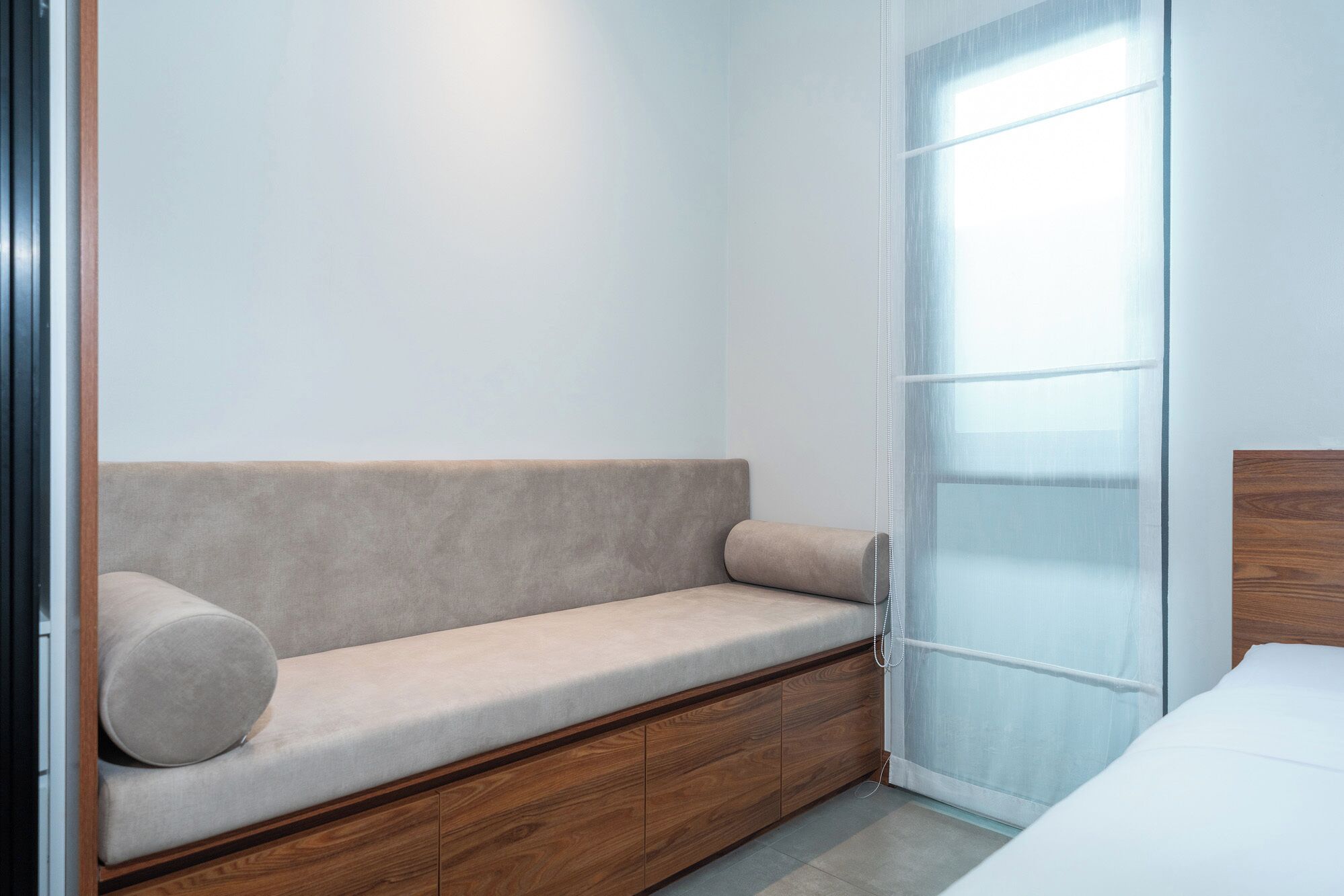 A minimalist room with a beige sofa against a white wall, a wooden cabinet, and a large window with sheer curtains.