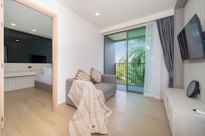 A modern living room with a sofa, a TV, and a balcony with a view of greenery. - Ozone 1Br Condo In Prestigious Laguna Area (Wichit)