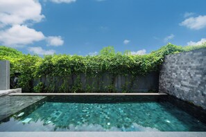 Uma piscina retangular com superfície de água azul translúcida, rodeada por um muro de pedra baixo e vegetação verde exuberante.