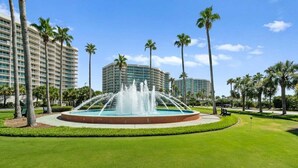 Un hotel con una fuente central, palmeras y un césped bien cuidado.