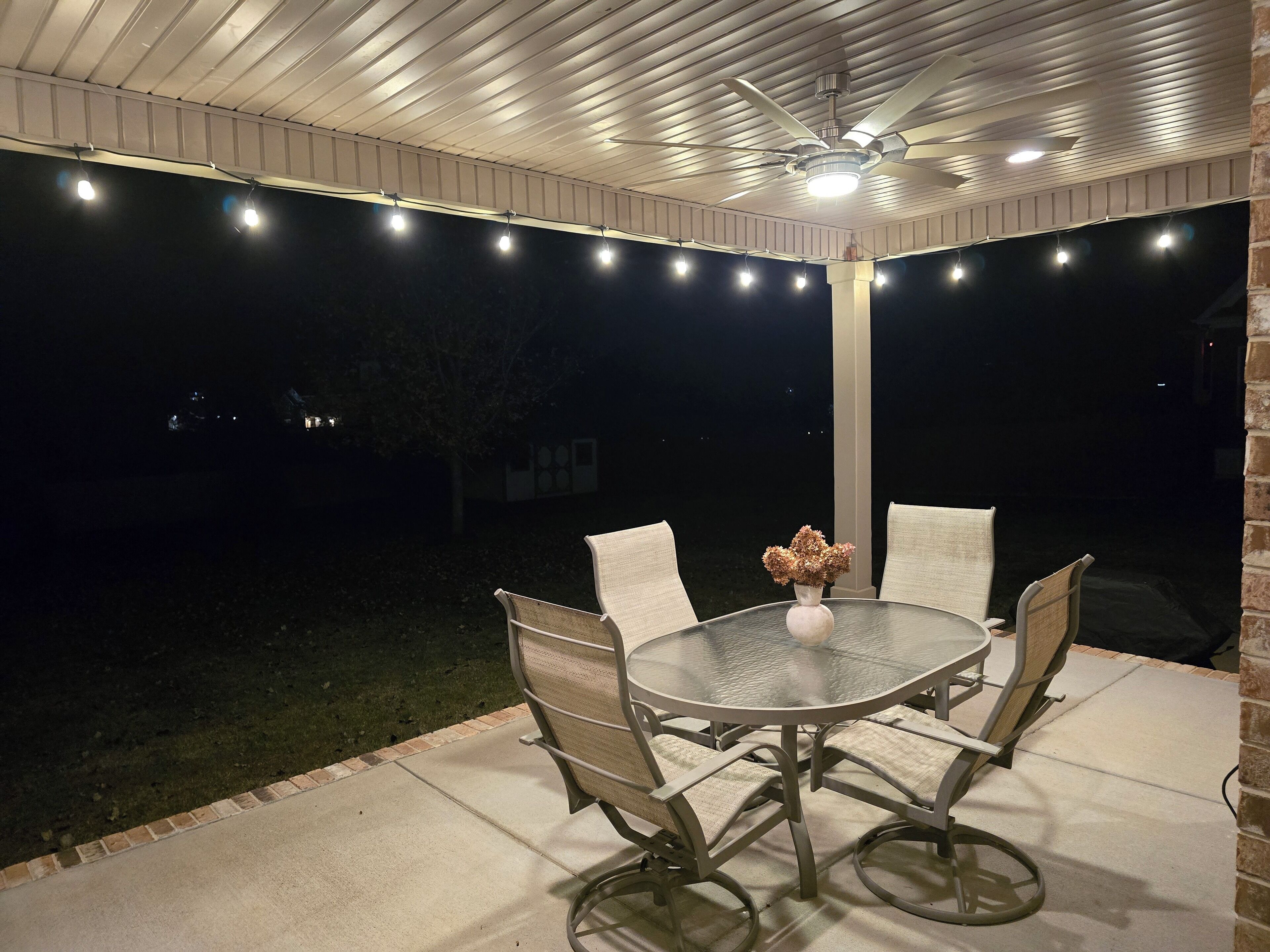 Eine Außenpatio mit einem runden Tisch, vier Stühlen, einem Deckenventilator und Lichterketten.