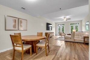 Amplia sala de estar con mesa de comedor de madera, asientos cómodos, televisor de pantalla plana y ventanas amplias con vistas a la vegetación.