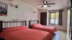 Una habitación con dos camas, un ventilador de techo, una ventana con cortinas y una lámpara fijada a la pared.