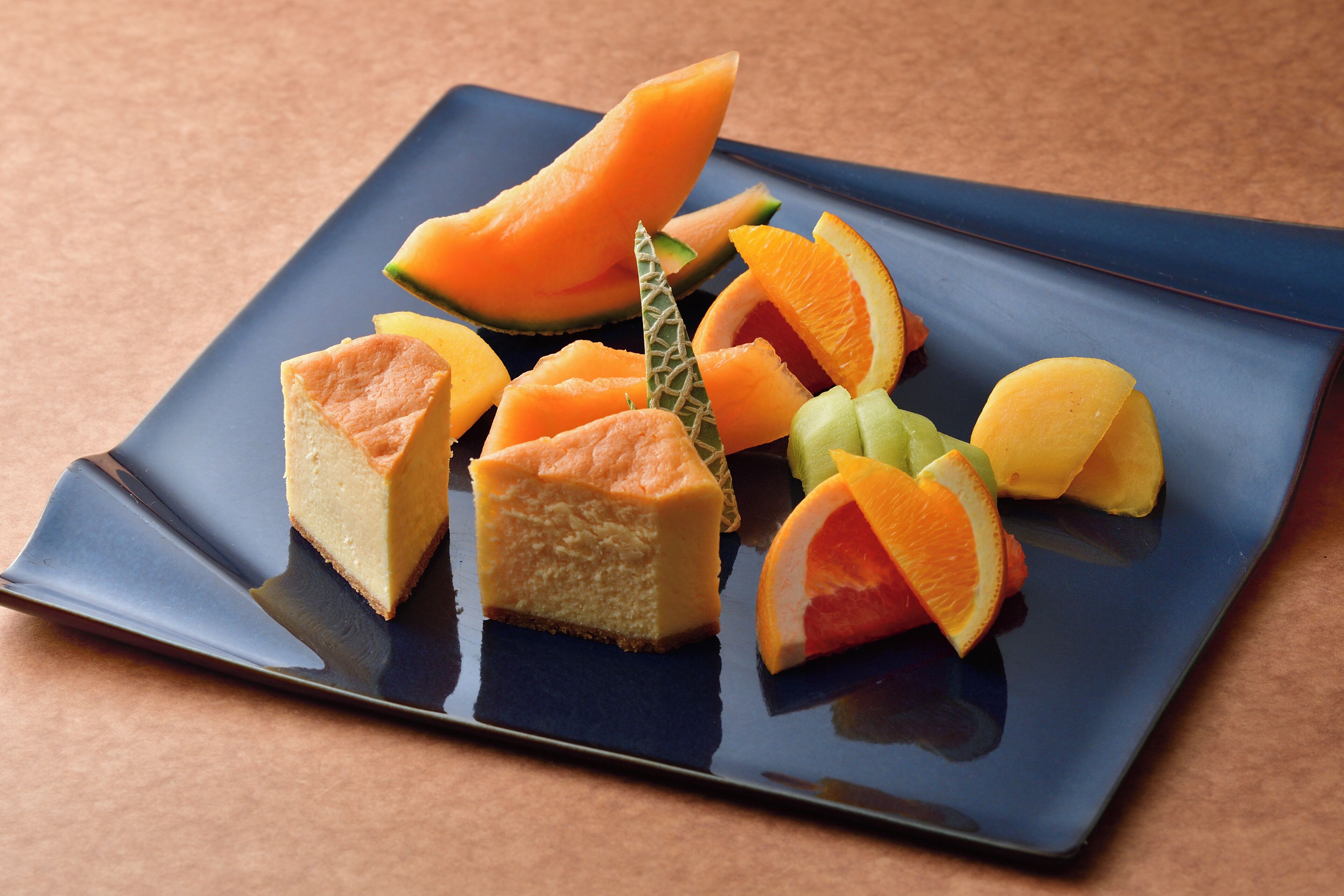 A square plate with various food items including melon slices, a cheesecake, and a decorative leaf.
