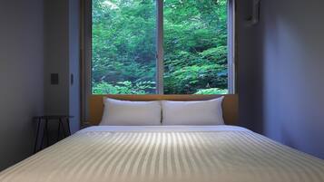 A bed with a wooden headboard and white pillows, a round table, and a window with a view of dense green foliage.