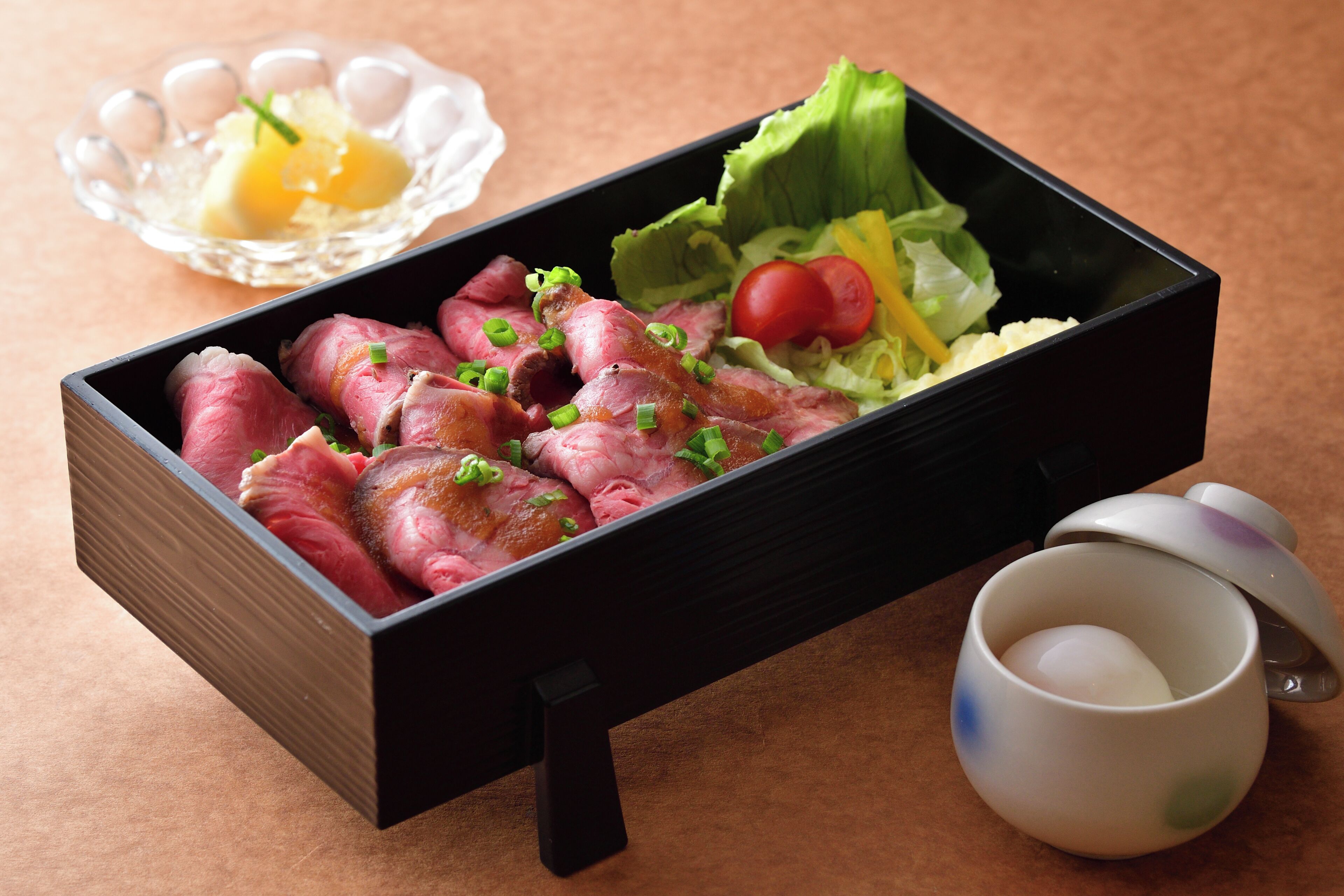 A rectangular black box containing sliced meat, garnished with green onions, and a side of lettuce and tomato.