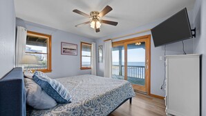 A bedroom with a bed, a ceiling fan, a TV, and a view of the ocean through a sliding glass door. - Sounds Good - Sound front home with stunning views and hot tub (Buxton)