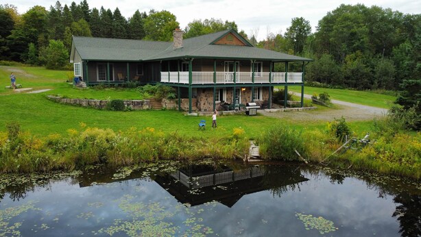 A lodge with a large porch, a stone chimney, and a pond with lily pads. - Cajun Cabin Private Nature Resort (Minden Hills)