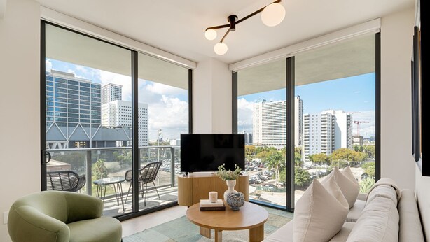 A modern living room with a sofa, armchair, and a flat-screen TV on a wooden stand. Large windows offer a city view. - Crosby Miami WTC 2BR/2BA Urban Comfort #614 (Miami)