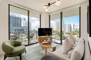 A modern living room with a sofa, armchair, and a flat-screen TV on a wooden stand. Large windows offer a city view. - Crosby Miami WTC 2BR/2BA Urban Comfort #614 (Miami)