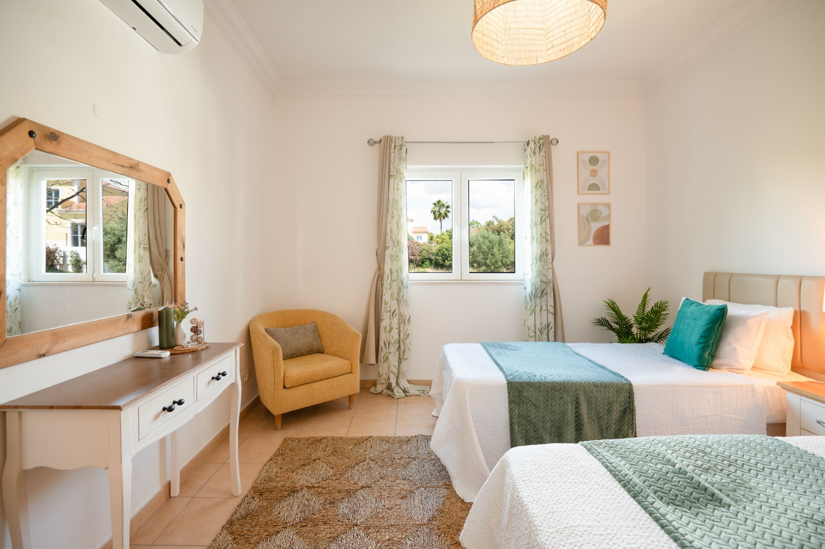 A bedroom with a wooden dresser, a chair, a bed with a green blanket, a bedside table with a lamp, and a window with curtains.