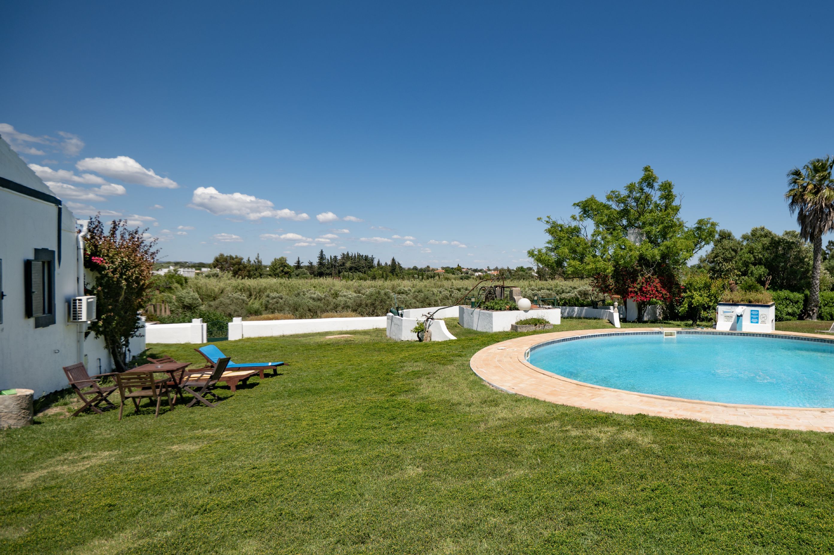 Une piscine entourée d’une pelouse, un bâtiment blanc et des arbres en arrière-plan.