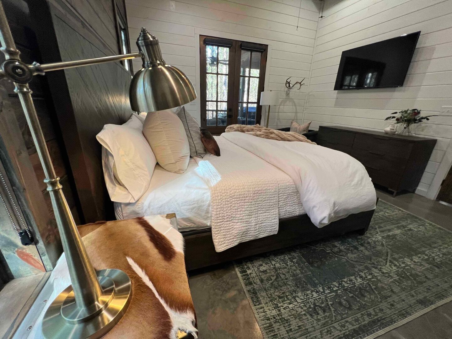 A bedroom with a bed, a desk lamp, a rug, and a television on the wall.