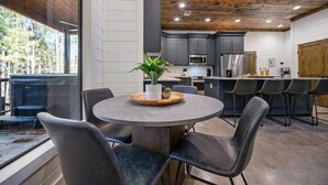 A modern kitchen with dark cabinets, a round dining table with chairs, and a large window overlooking a forest. - Swing, Games, EV, Hot Tub, Fire Pit, Black Diamond (Broken Bow)