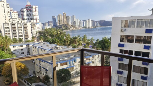 A balcony with red chairs and a glass table overlooking a cityscape and a body of water. - APARTMENT IN SANTA MARTA EL RODADERO LAURELES BUILDING 801 With ocean view (Gaira)