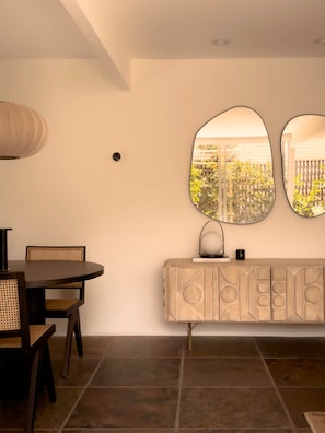 A dining area with a round table, chairs, a cabinet with geometric patterns, and two wall-mounted mirrors reflecting a garden view. - Historic 1BR 2BR Retreat w Outdoor Shower & Private Patio in Palm Springs (Palm Springs)