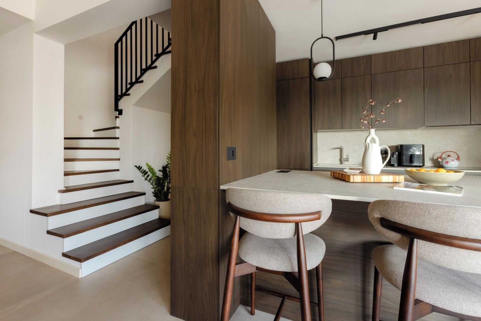 A modern kitchen with a central island, bar stools, a staircase, and wooden cabinetry.