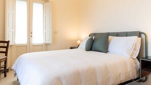 Ein ordentlich bezogenes Bett mit weißen Laken, ein Nachttisch mit Lampe und ein Sessel.