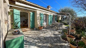 Une cour dallée de pierres, avec un espace repas, des volets verts et des plantes en pot.