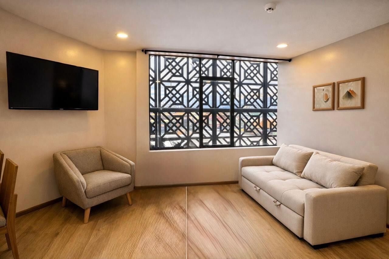 A modern living room with a flat-screen TV, a beige sofa, a wooden chair, and a large window with a geometric pattern.