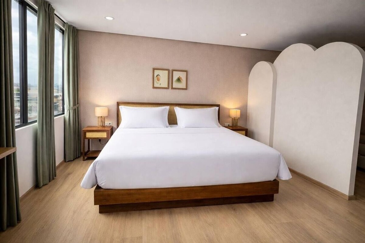 Una cama grande con ropa de cama blanca, dos mesitas de noche con l ámparas, vistas de la ciudad a través de amplias ventanas y un panel decorativo en la pared.