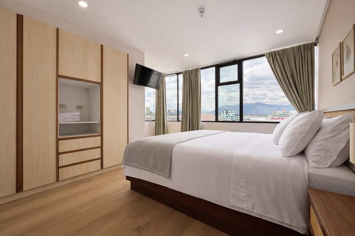 A hotel room with a bed, a TV, a wardrobe, and a window with a view of buildings and mountains.