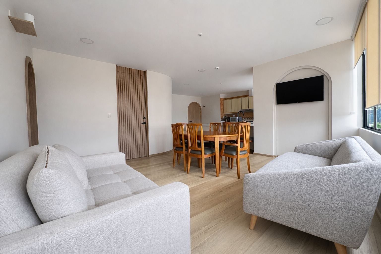 Ein modernes Wohnzimmer mit zwei grauen Sofas, einem Holzesstisch mit Stühlen, einer Küche im Hintergrund und einem an der Wand montierten Fernseher.