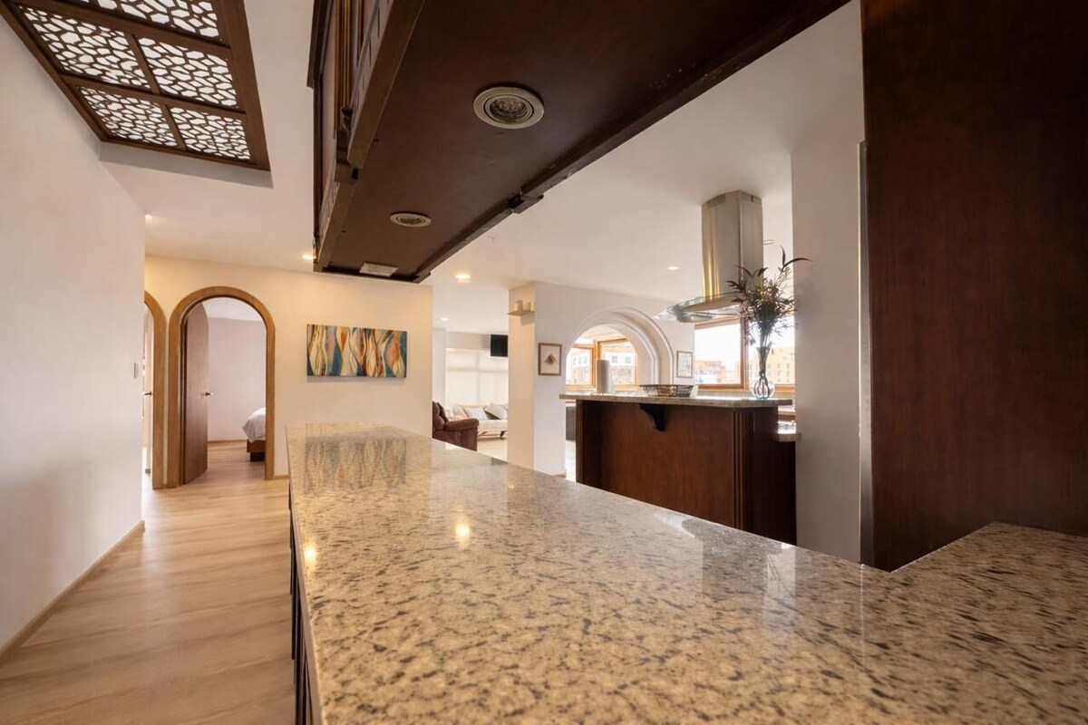 Una cocina moderna con encimeras de granito, techo de madera oscura y vistas al salón.
