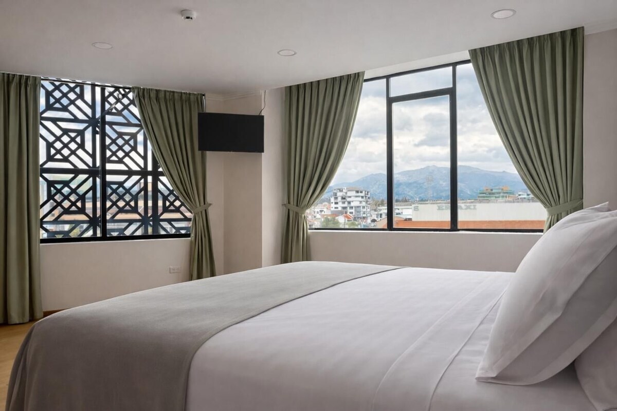Habitación de hotel con una cama, un ventanal con cortinas y vistas a edificios y montañas.
