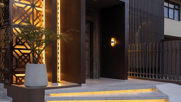 Una entrada moderna a un edificio con fachada geométrica, un amplio arco de acceso y una escalera con peldaños iluminados.