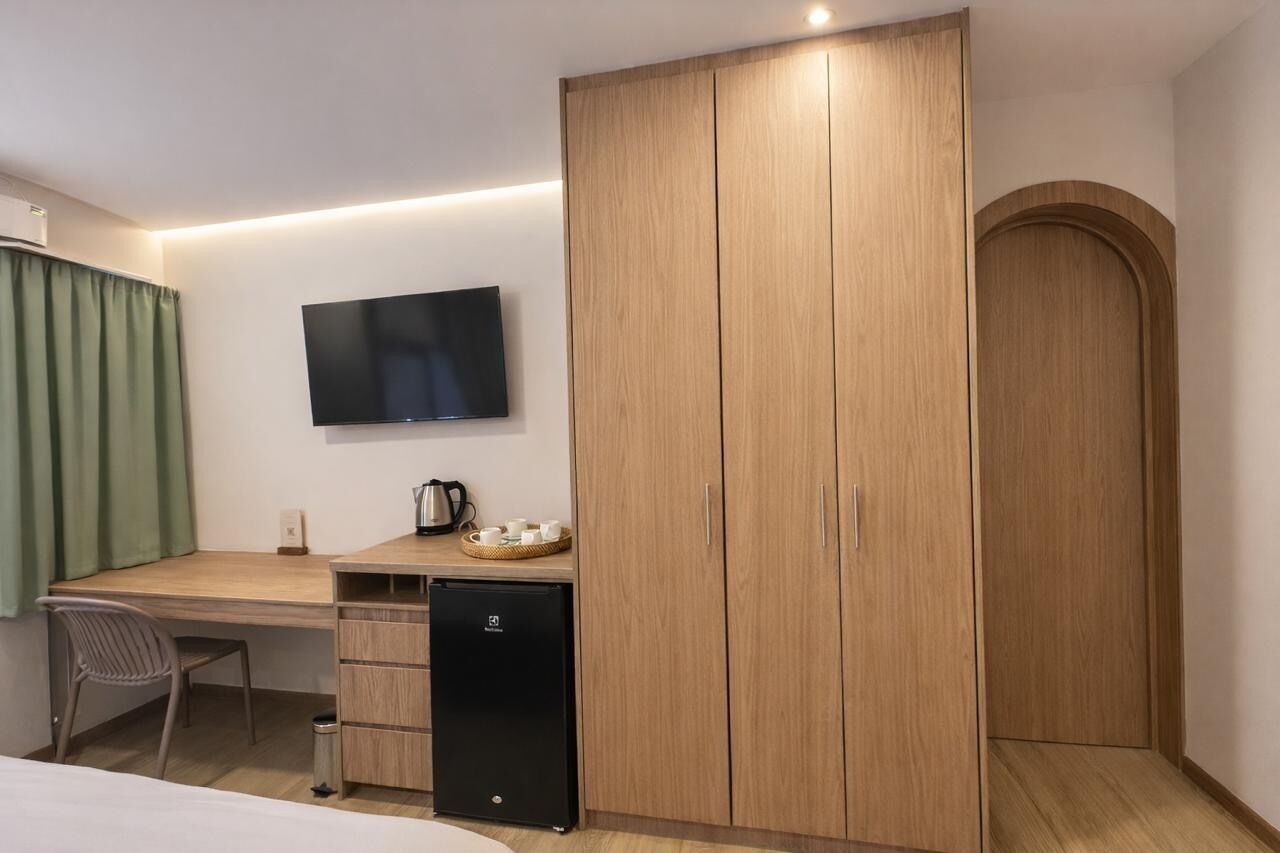 A hotel room with a wooden wardrobe, a desk with a television, a small refrigerator, and a green curtain.
