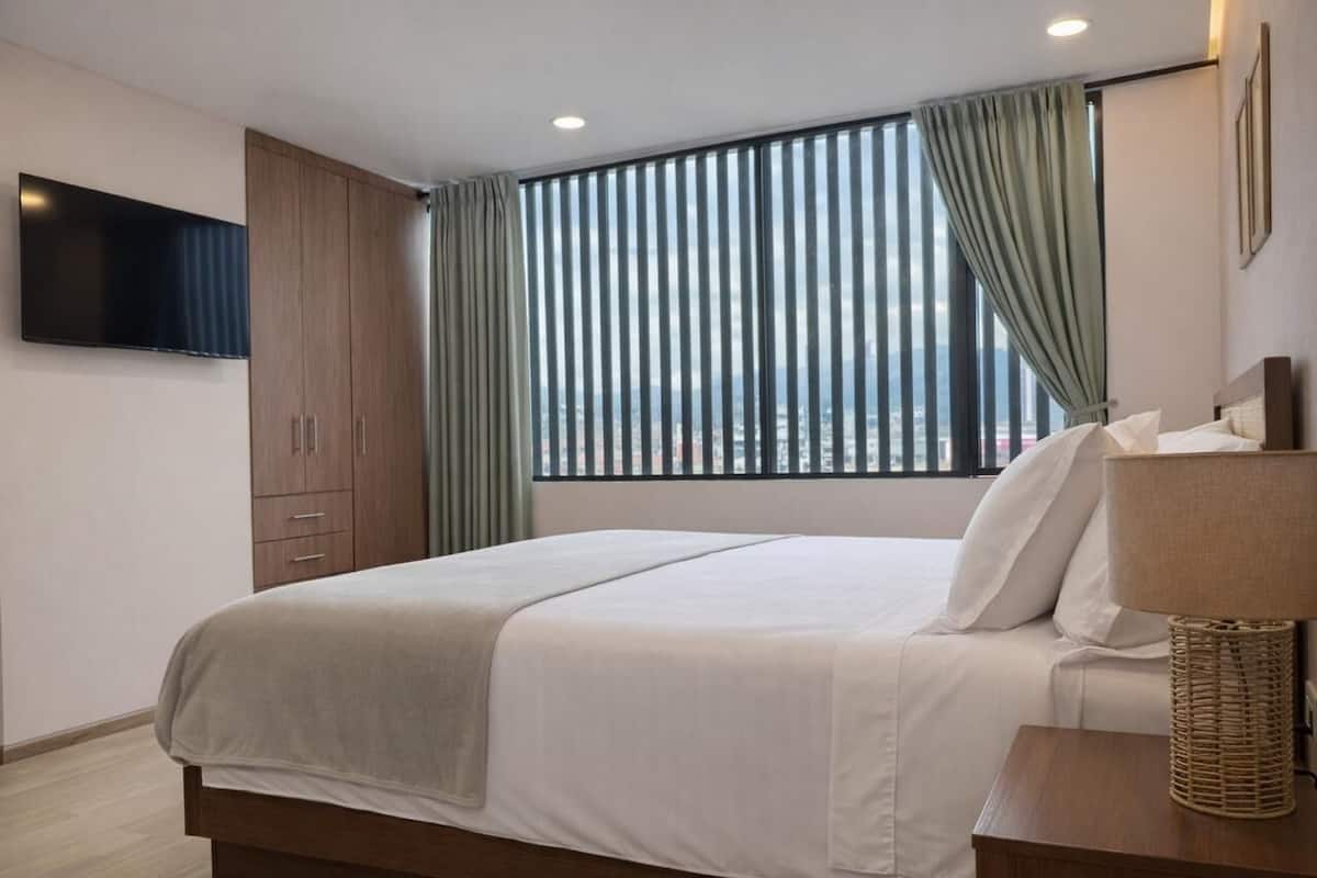 A modern hotel room with a large bed, a flat-screen TV mounted on the wall, and a view of the city through floor-to-ceiling windows.