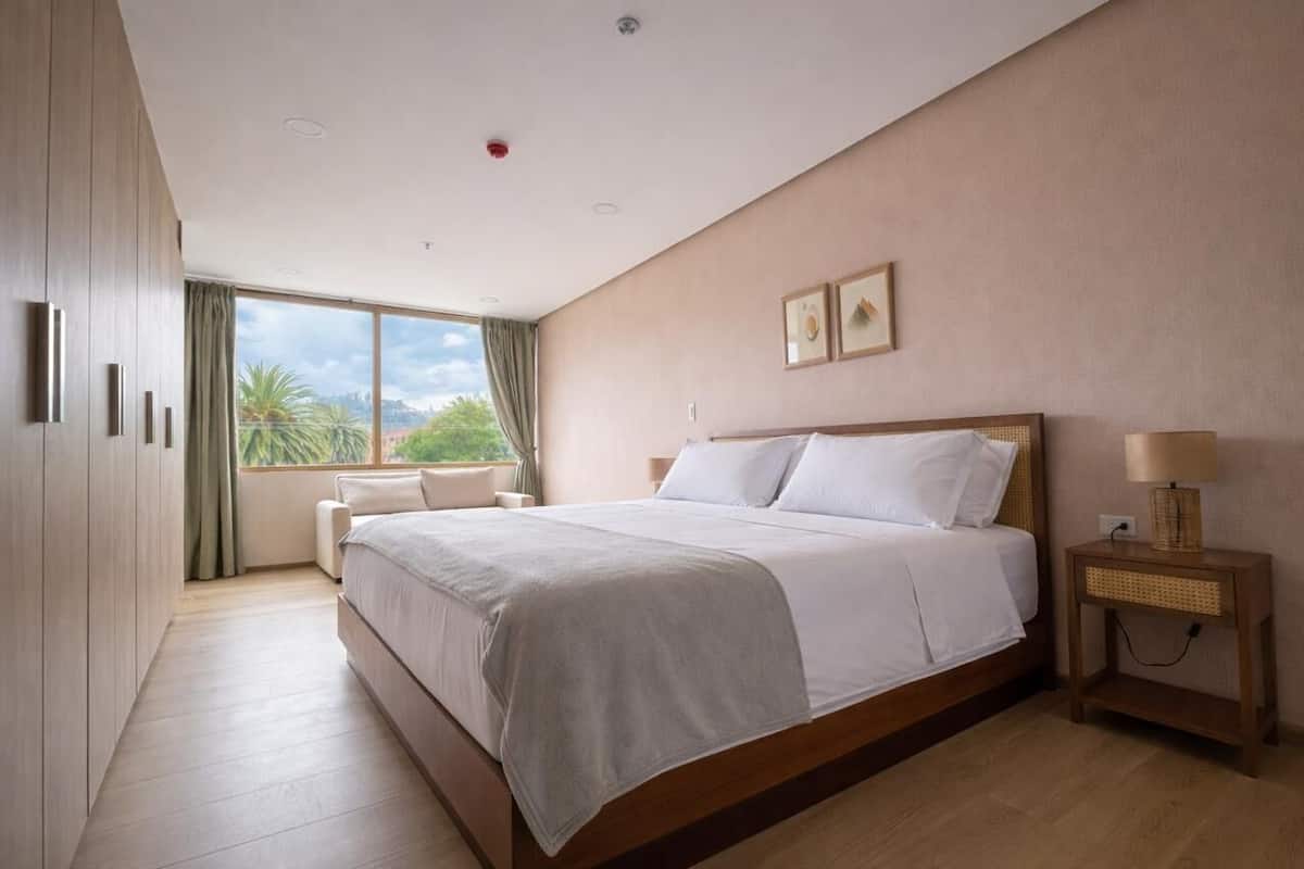 A bedroom with a large bed, a nightstand, a wardrobe, and a window with a view of greenery.