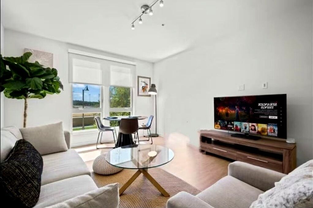 A modern living room with a glass coffee table, a sofa, a TV on a wooden stand, and a large window offering a view of the outdoors.