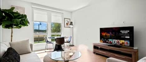 A modern living room with a glass coffee table, a sofa, a TV on a wooden stand, and a large window offering a view of the outdoors.
