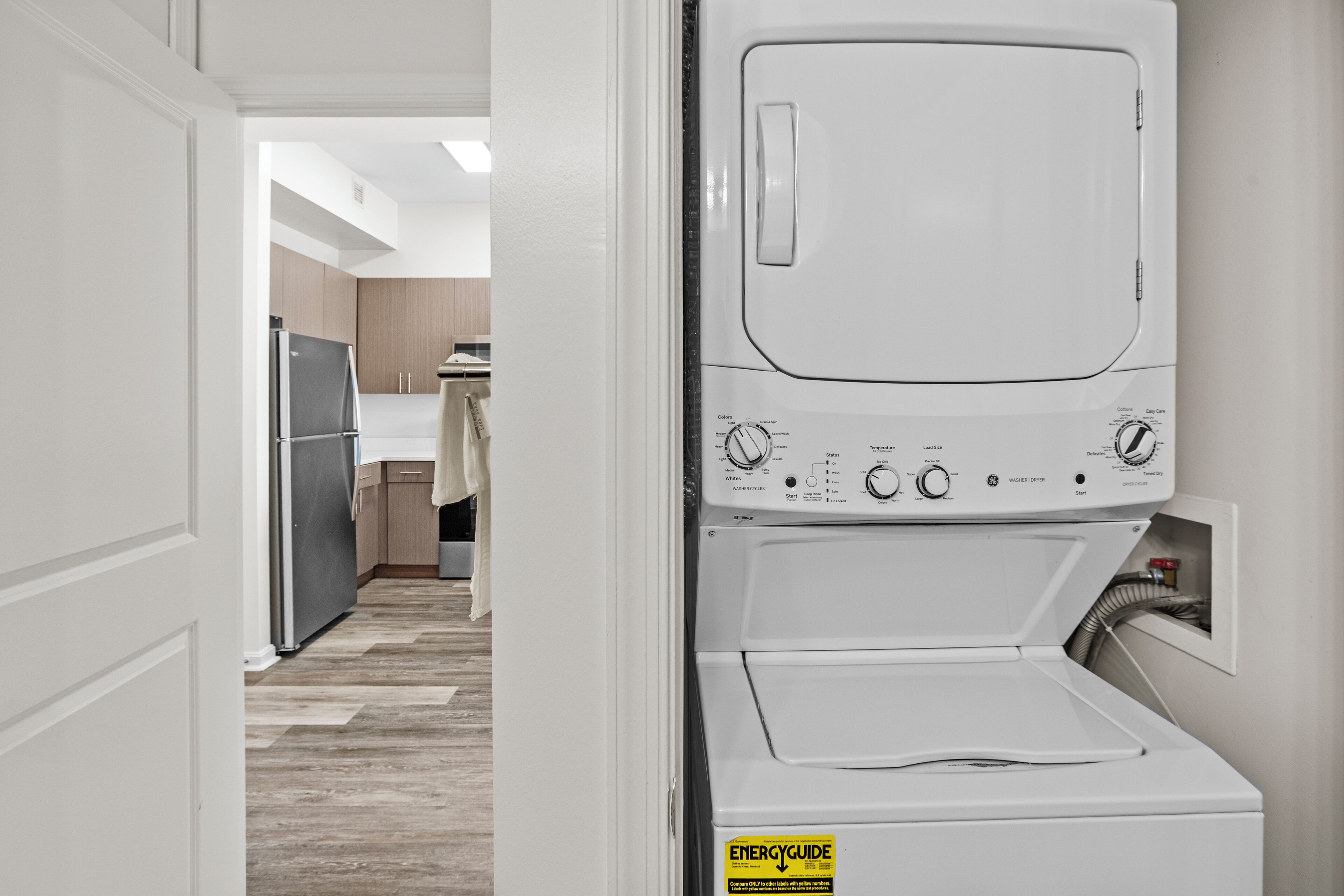 A laundry room with a front-loading washer and dryer, a wooden floor, and a kitchen area in the background.