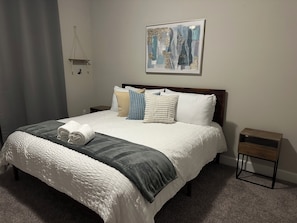 A neatly made bed with a white comforter, a gray throw blanket, and multiple pillows. There is a small bedside table with a lamp and a framed abstract painting above the bed. - Spacious 5-bedroom house in beautiful Loughman with AC and gym (Loughman)