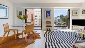 A modern living room with a dining area, a large window with a city view, and a flat-screen TV. - Designer Sub-Penthouse in Potts Point (Elizabeth Bay)