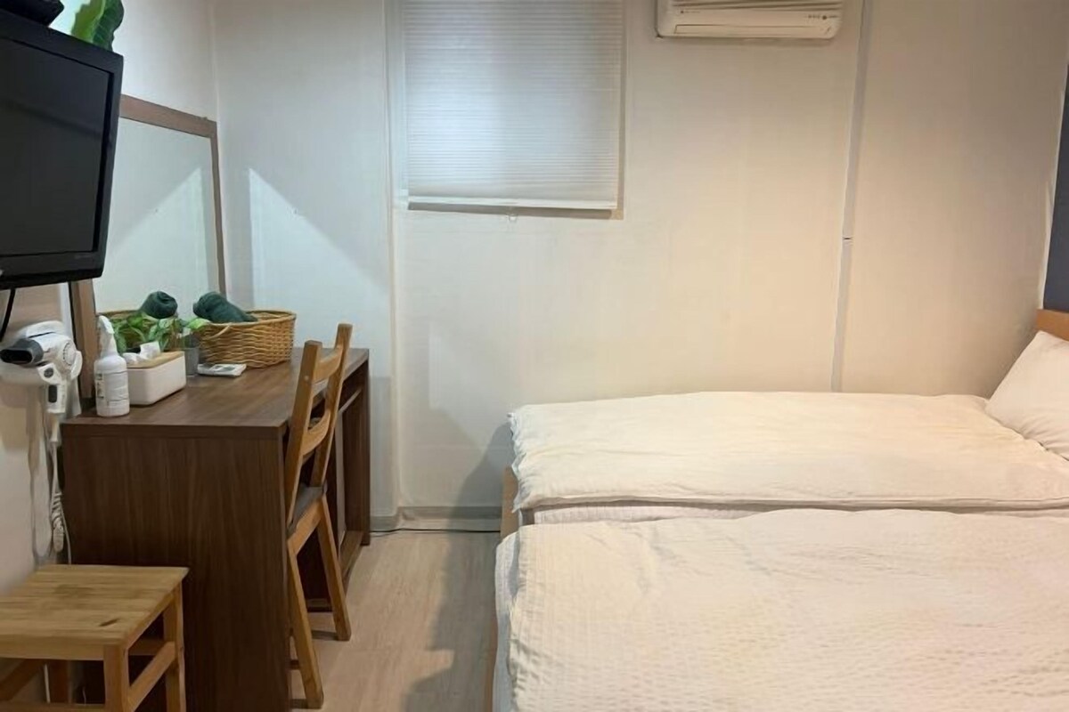 A bedroom with a bed, a desk with a chair, a television, and a window with blinds.