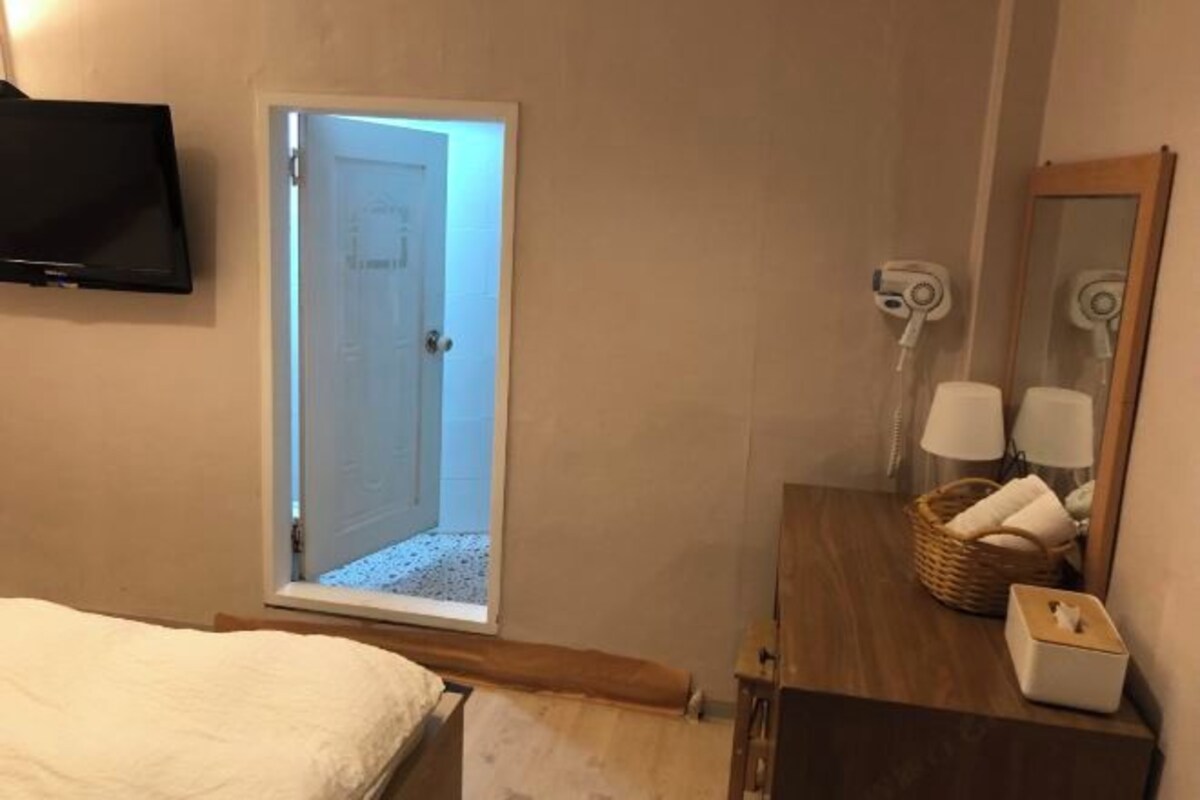 A bedroom with a bed, a TV, a bathroom with a mirror, and a door leading to a hallway.
