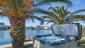 A hot tub with a wooden deck and a palm tree in the foreground, a body of water, and houses in the background. - Waterfront Canal Hot Tub Kayaks (Hernando Beach)