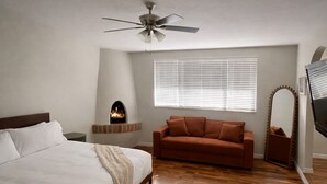 Une chambre à coucher comprenant un lit, un canapé, une cheminée, un ventilateur de plafond et une fenêtre avec des stores.