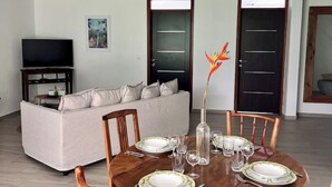 A dining area with a round wooden table set for four, a sofa, and a television in the background. - Ti Bwa Lodge - Garden villa with pool and sea view (Capesterre-Belle-Eau)