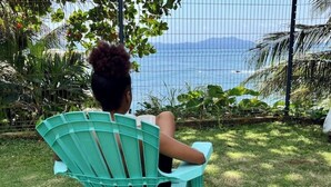 A person seated on a green chair facing a body of water, with a fence and tropical plants in the background. - Ti Bwa Lodge - Garden villa with pool and sea view (Capesterre-Belle-Eau)