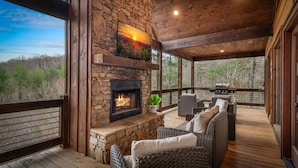 A covered outdoor area with a stone fireplace, wicker furniture, and a view of the surrounding forest. - 249 Ranger Station - Mountain View | Pet Friendly | Hot Tub | Shuffleboard | Cornhole (Morganton)
