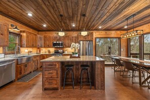 A rustic kitchen with wooden cabinets, a central island with stools, stainless steel appliances, and large windows. - 249 Ranger Station - Mountain View | Pet Friendly | Hot Tub | Shuffleboard | Cornhole (Morganton)