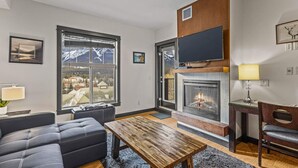 A modern living room with a fireplace, large windows, and a cozy seating area. - Relaxing 1BR Getaway. Hot tub + Amazing Mtn Views (Canmore)