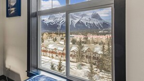 A room with a large window offering a view of snow-covered mountains and a town below. - Relaxing 1BR Getaway. Hot tub + Amazing Mtn Views (Canmore)
