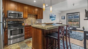 A modern kitchen with wooden cabinets, stainless steel appliances, a granite countertop, and a large island with bar stools. - Relaxing 1BR Getaway. Hot tub + Amazing Mtn Views (Canmore)